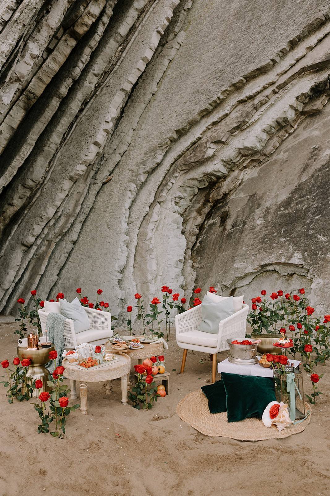 A styled beach setting — armchairs and dessert tables among long-stemmed roses, against the Basque cliffs