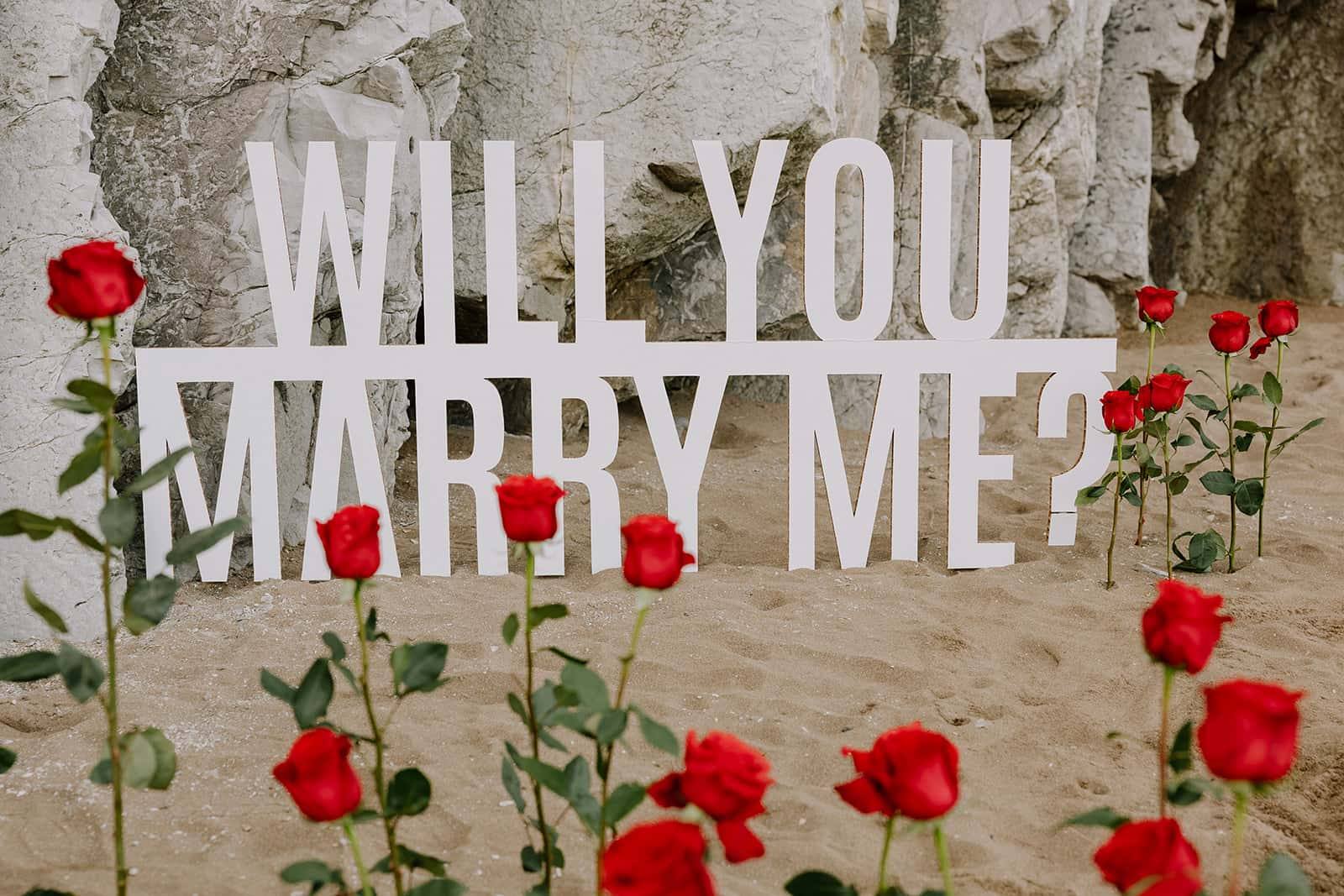 A large “Will you marry me?” sign among red roses on the sand