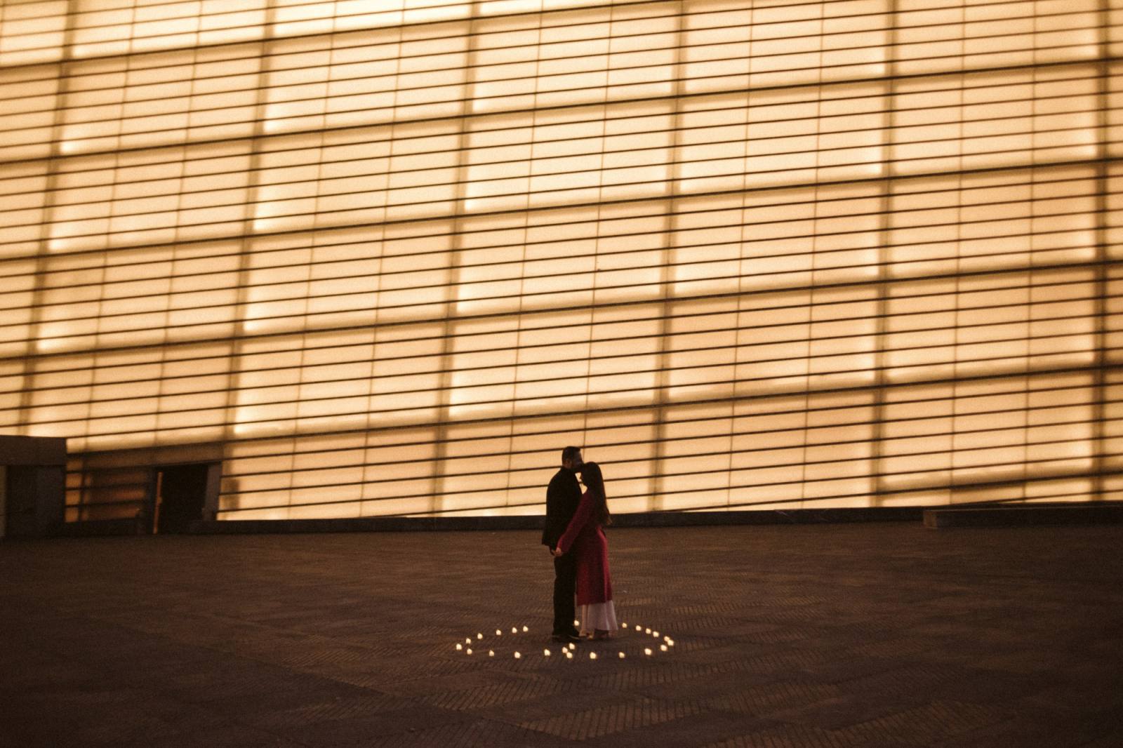 A couple embracing inside a heart of candles before the glowing glass facade of the Kursaal, San Sebastián, at dusk